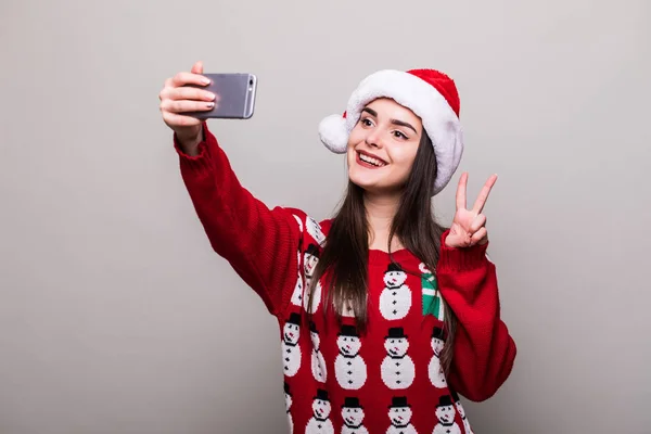 Menina modelo desgaste santa chapéu e Natal suéter tomar selfie no fundo cinza — Fotografia de Stock