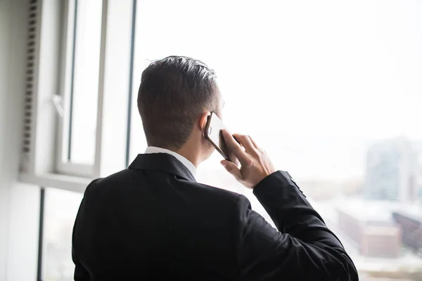 Pohledný mladý podnikatel stojící vedle velkými okny své kanceláře nejvyššího patra, když mluví na svůj mobilní telefon při pohledu na pohled na město — Stock fotografie