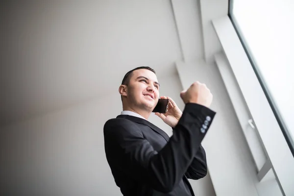 Jeune homme d'affaires debout à côté de grandes fenêtres de son bureau au dernier étage, regardant la vue sur la ville tout en prenant de bonnes nouvelles de son discours sur son téléphone portable — Photo