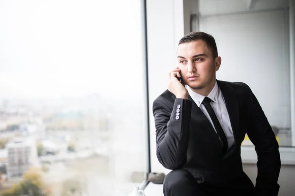 Hombre de negocios en un elegante espacio de oficina moderno con grandes ventanales, hablando por teléfono y mirando hacia el exterior — Foto de Stock