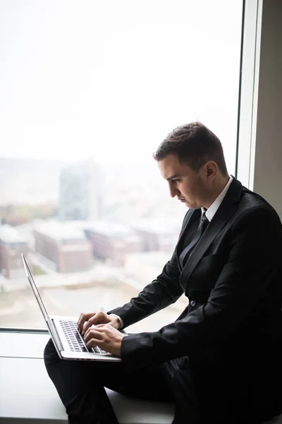 Giovane uomo d'affari che lavora vicino alla finestra panoramica della città sul computer portatile — Foto Stock