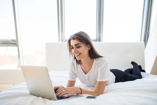 Retrato de una hermosa joven feliz tumbada en la cama y usando el ordenador portátil en casa — Foto de Stock