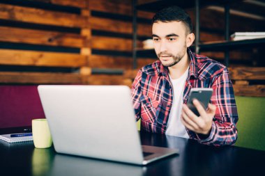 telefon ve office içinde laptop ile tablo başına oturmuş tişörtlü adam.