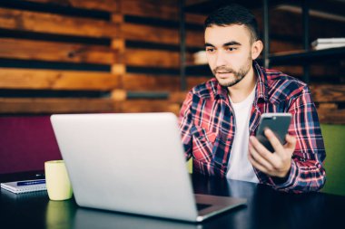 telefon ve office içinde laptop ile tablo başına oturmuş tişörtlü adam.