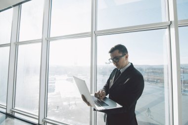 Takım elbise ve gözlük çalışmalarında yakışıklı işadamı ile laptop ofis panoramik pencerelere karşı elinde