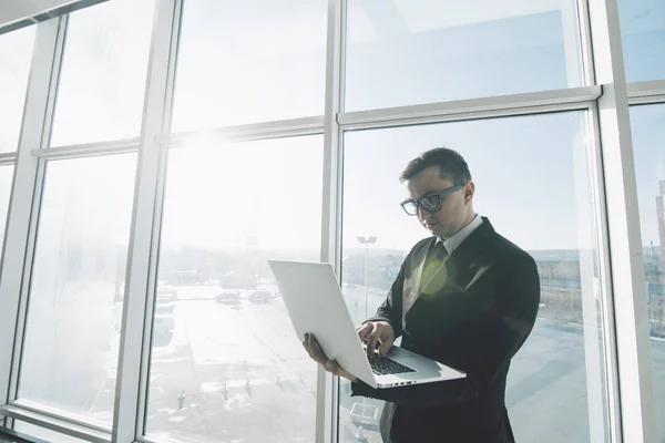 Beau businessman en costume et lunettes de travail avec ordinateur portable dans les mains dans le bureau contre les fenêtres panoramiques — Photo