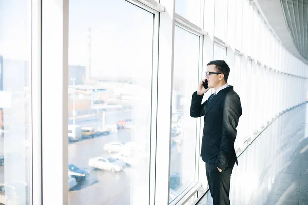 Beau homme d'affaires debout à côté des grandes fenêtres de son bureau au dernier étage, regardant la vue sur la ville tout en parlant sur son téléphone portable — Photo