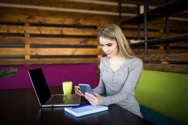 Cep telefonu, dizüstü bilgisayar, kupa ve defter bakışla masada oturan genç kadın. Coworking.