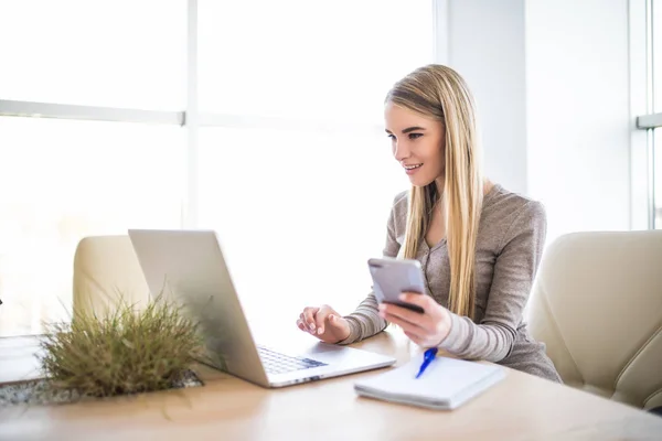Portret van een jonge zakenvrouw met telefoon in de hand via de laptop op kantoor — Stockfoto