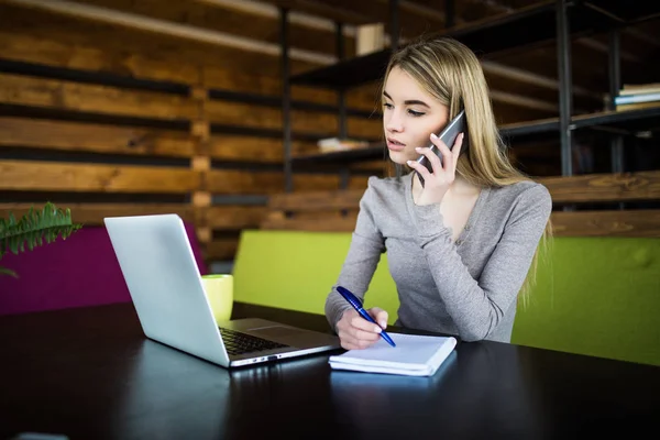 Joven mujer de negocios en coworking hablar por teléfono y hacer notar en el cuaderno — Foto de Stock