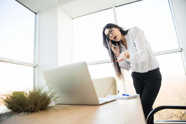 Επιχειρήσεων γυναίκα μιλάει στο τηλέφωνο και να εξετάσουμε το φορητό υπολογιστή στο γραφείο της. — Φωτογραφία Αρχείου