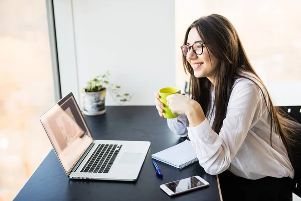 Junge Arbeiterin sitzt im Büro, während sie Laptop benutzt und Kaffee trinkt — Stockfoto