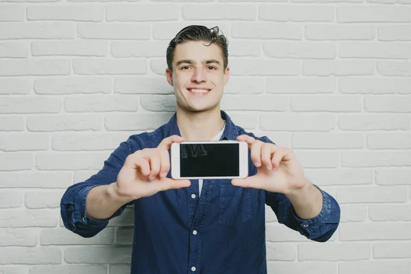 Hermoso joven en ropa casual mostrar teléfono con pantalla negra sobre un fondo de pared de ladrillo blanco —  Fotos de Stock