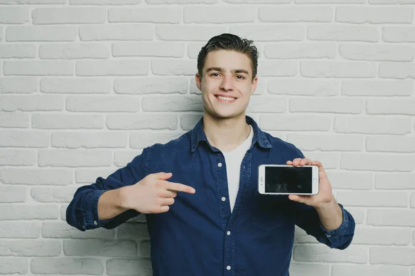 Retrato de joven empresario atractivo en ropa casual señaló un teléfono inteligente, mirando en cámara y sonriendo, de pie contra la pared de ladrillo — Foto de Stock