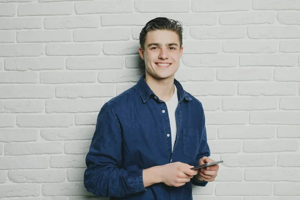 ハンサムな若い男の携帯電話を保持しているとレンガの壁に立っている間それを見て — ストック写真