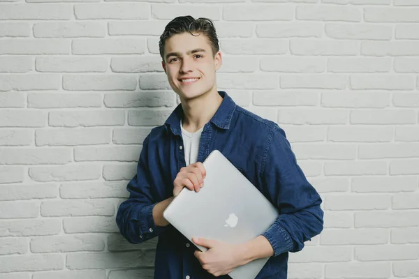 Schöner Mann lächelt, während er Laptop in der Hand hält und gegen Ziegelmauer steht — Stockfoto