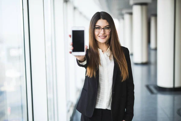 Attraente donna d'affari in possesso di uno smartphone, che punta su di esso, guardando la fotocamera e sorridendo . — Foto Stock
