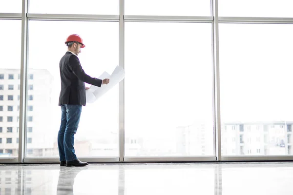 Builder ingenjör slitage säkerhet hjälm titt på blueprint papper konstruktion nära panoramafönster — Stockfoto