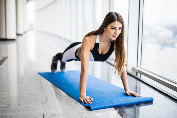 Working her core muscles. Full length of young beautiful woman in sportswear doing plank while standing in front of window at gym — Stock Photo, Image