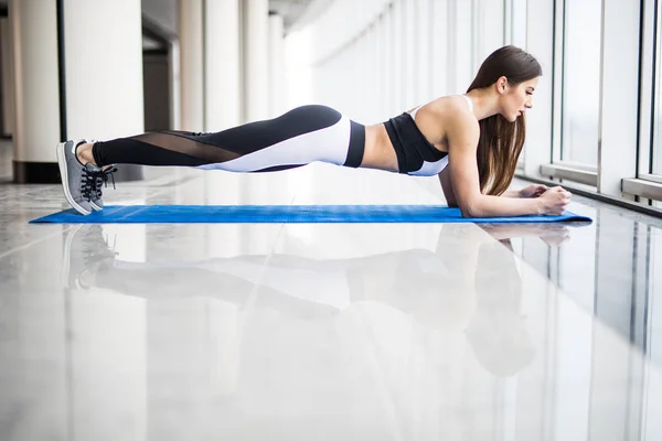 Vista lateral de larga duración de la joven hermosa mujer en ropa deportiva haciendo tablón mientras está de pie frente a la ventana en el gimnasio — Foto de Stock