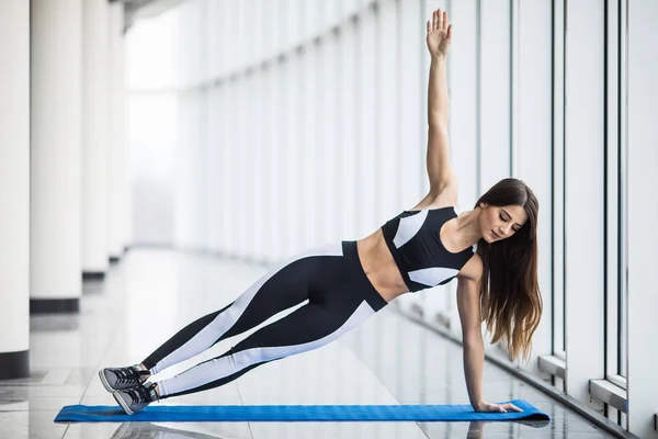 Panjang penuh wanita cantik muda dalam pakaian olahraga melakukan sisi papan di depan jendela di gym — Stok Foto