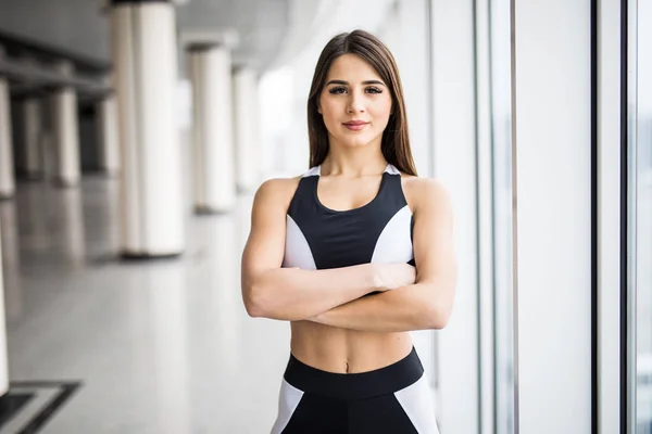 Ung kvinna med korsade händer i sportkläder tittar kameran stående framför fönstret på gym — Stockfoto