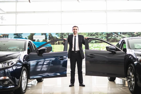 Confident smiling car salesman at the showroom near two cars, he is standing near open cars doors of luxury cars