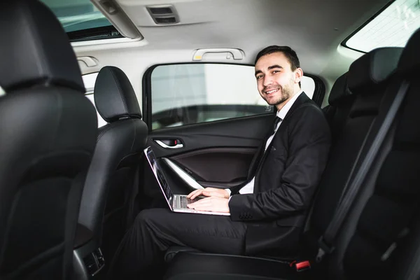 Schöner junger Geschäftsmann arbeitet an seinem Laptop, während er im Auto sitzt und in die Kamera schaut — Stockfoto