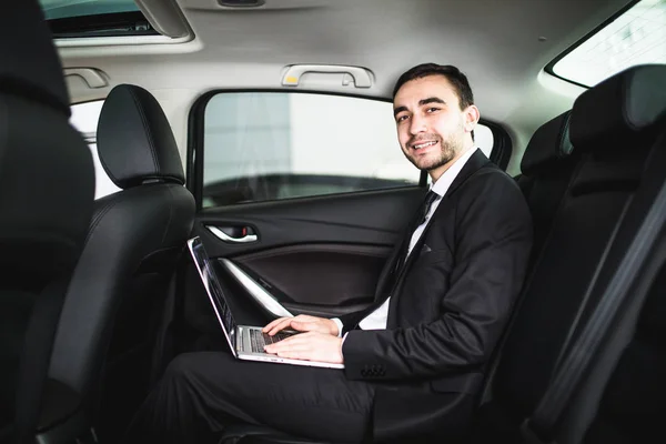 Schöner junger Geschäftsmann arbeitet an seinem Laptop, während er im Auto sitzt und in die Kamera schaut — Stockfoto