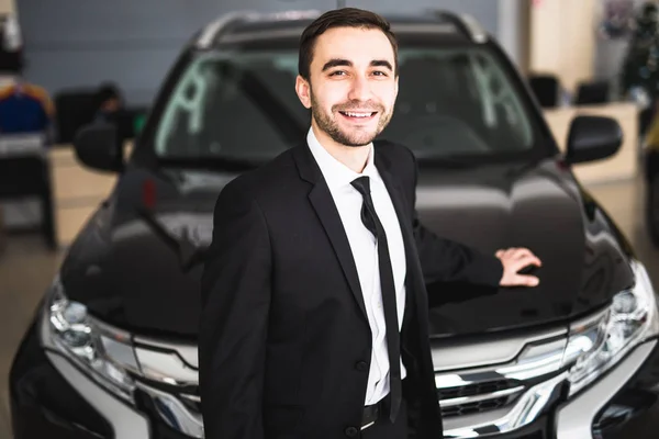 Bonito jovem vendedor de carros clássico em pé na concessionária na frente de carro novo — Fotografia de Stock