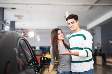 Güzel genç çift ayakta araba satın almak seçme galerisinde. Adam arabanın üzerine işaret.