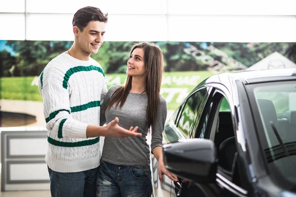 Belo jovem casal de pé na concessionária escolhendo o carro para comprar. Homem apontado no carro . — Fotografia de Stock