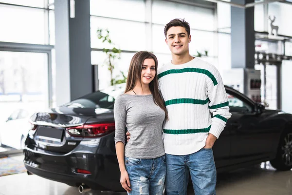 Sorrindo casal de pé na frente de um carro no showroom do carro novo — Fotografia de Stock