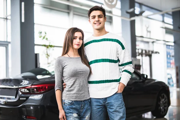 Sorrindo casal de pé na frente de um carro no showroom do carro novo — Fotografia de Stock