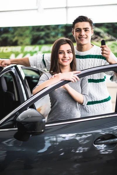 Bela jovem casal está sorrindo e olhando para a câmera enquanto se inclina em seu novo carro em um show de motor. Homem está segurando chaves do carro — Fotografia de Stock