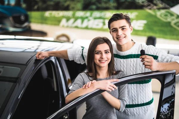 Bela jovem casal está sorrindo e olhando para a câmera enquanto se inclina em seu novo carro em um show de motor. Homem está segurando chaves do carro — Fotografia de Stock