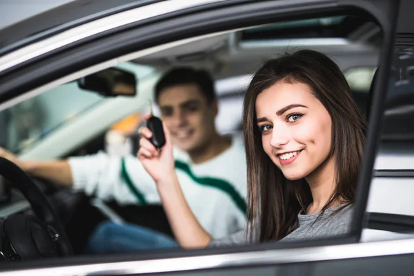 Belo jovem casal sentado nos bancos da frente de seu novo carro, enquanto a mulher mostrando chaves e sorrindo — Fotografia de Stock