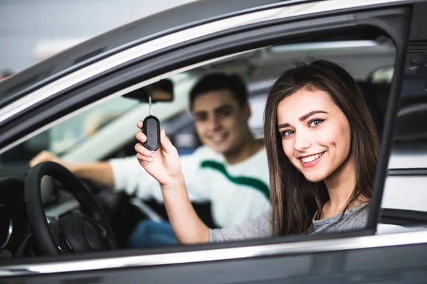 Belo jovem casal sentado nos bancos da frente de seu novo carro, enquanto a mulher mostrando chaves e sorrindo — Fotografia de Stock