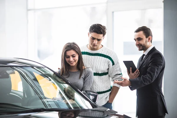 Beau jeune couple choisissant une voiture chez le concessionnaire parlant au gestionnaire de salon avec tablette dans les mains — Photo