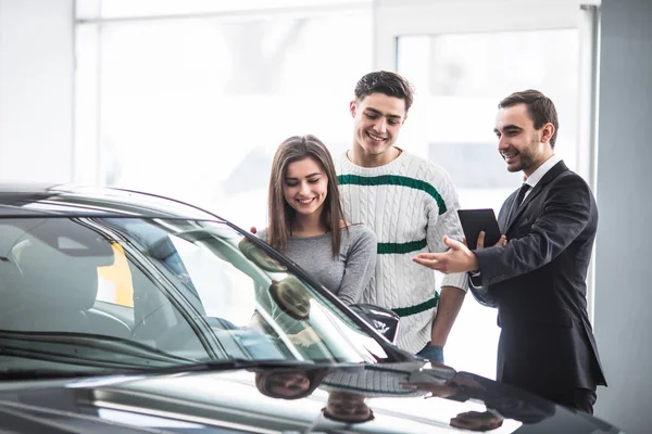 Beautiful young couple choosing a car at the dealership talking to the salon manager with tablet in hands — Stock Photo, Image