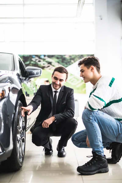 Young car salesman showing the advantages of the car to the customer and tires. — Stock Photo, Image