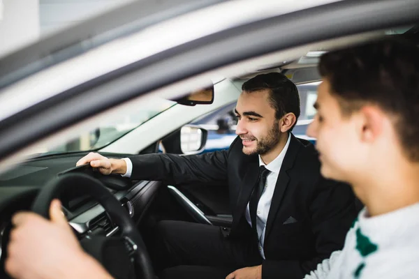 Let me show you interior of this car. Handsome young classic car salesman standing in the dealership and helping a client to make a decision about new car — Stock Photo, Image