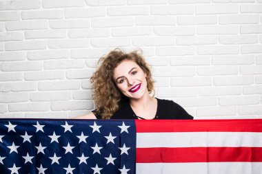 Güzel genç kıvırcık kız poz ve gülümseyen rahat kıyafetler, ayakta duvara karşı Amerikan bayrağı ile kaplı