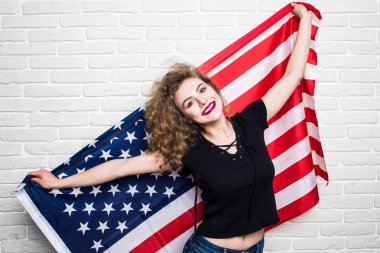 Güzel genç kıvırcık kız poz ve gülümseyen rahat kıyafetler, ayakta duvara karşı Amerikan bayrağı ile kaplı