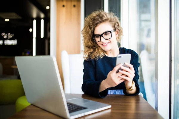 Mujer joven y atractiva revisando sus mensajes de texto en su teléfono móvil con una sonrisa mientras se sienta en su escritorio en la oficina. Mira el portátil — Foto de Stock