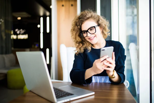 Attraktiv ung kvinna kolla hennes textmeddelanden på sin mobiltelefon med ett leende som hon sitter vid sitt skrivbord på kontoret. Titta på laptop — Stockfoto