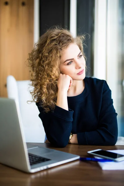 Retrato de una mujer joven pensando en algo mientras está sentada frente a la computadora portátil portátil en el interior moderno . — Foto de Stock