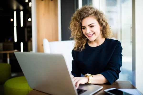 Gelukkig curlu jonge vrouw op moderne plaats met heldere windows laptop gebruikt. — Stockfoto