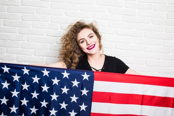 Красивая молодая кудрявая девушка в повседневной одежде позирует и улыбается, стоя покрытая американским флагом на кирпичной стене — стоковое фото