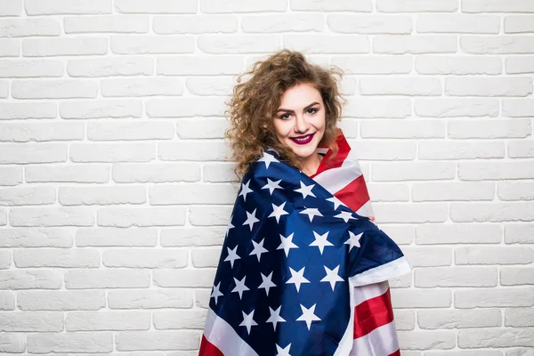 Красивая молодая кудрявая девушка в повседневной одежде позирует и улыбается, стоя покрытая американским флагом на кирпичной стене — стоковое фото